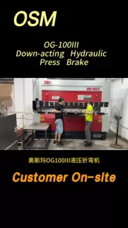 OSM OG-100III down-acting hydraulic press brake in operation. This industrial metal folding machine features a robust red and white design, shown here with a customer on-site testing the equipment for precision bending applications.